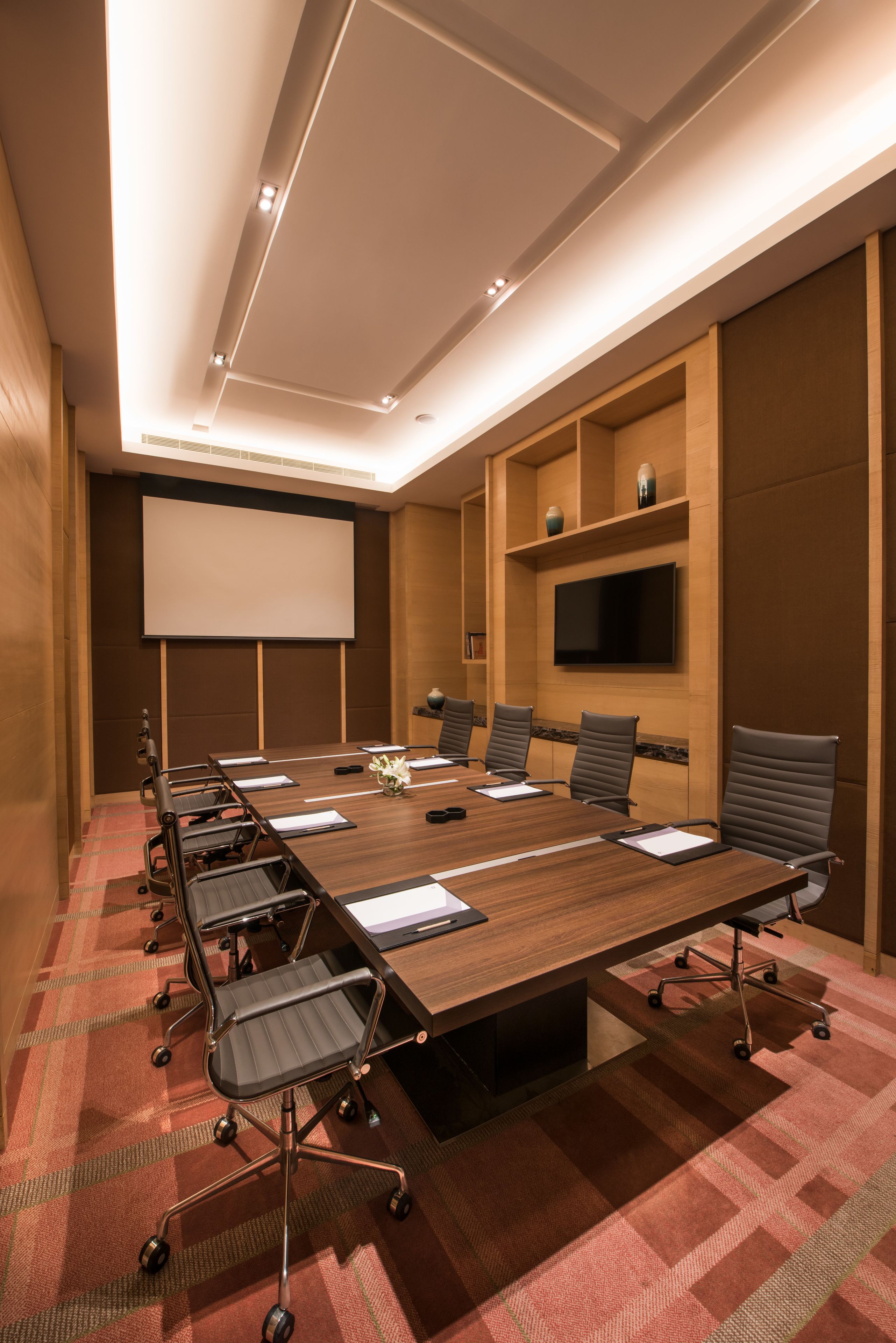 Sala de conferência com uma mesa comprida, cadeiras, uma tela de projetor e uma televisão de tela plana na parede.