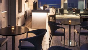 An outdoor dining area with round tables and wicker chairs, a bar area with stools, and a view of the cityscape at dusk. - Hyatt Regency Lucknow (Lucknow)