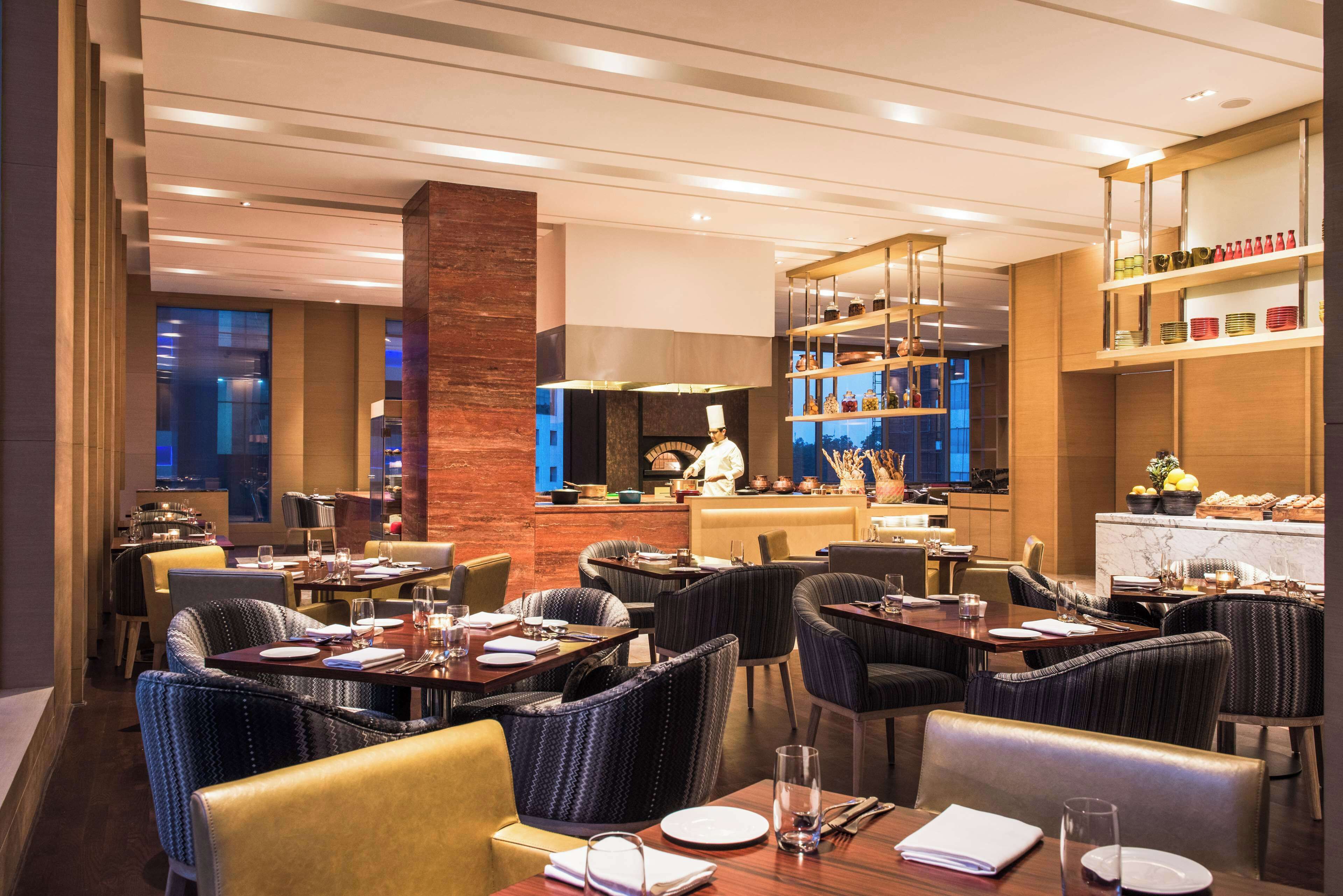 A modern restaurant with a chef preparing food, dining tables with chairs, and a bar area.