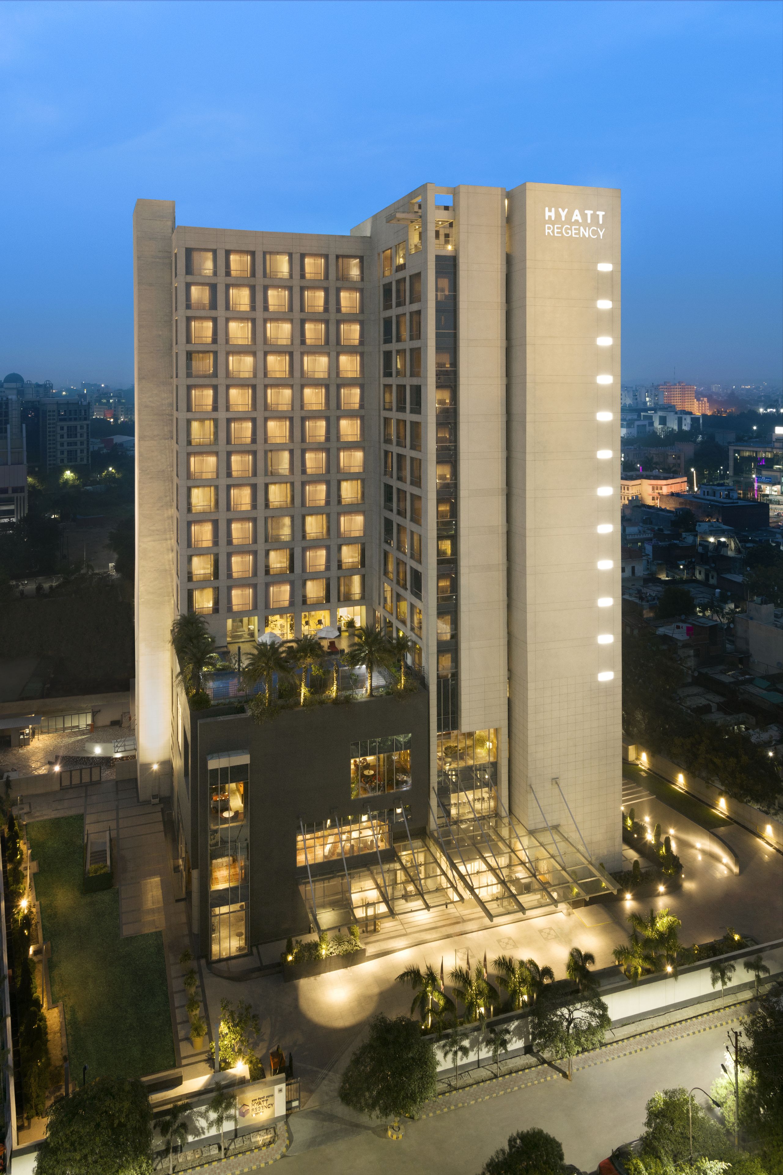 Um hotel moderno de vários andares, com uma placa chamativa de "HYATT REGENCY", diversas varandas e uma área de entrada bem iluminada.