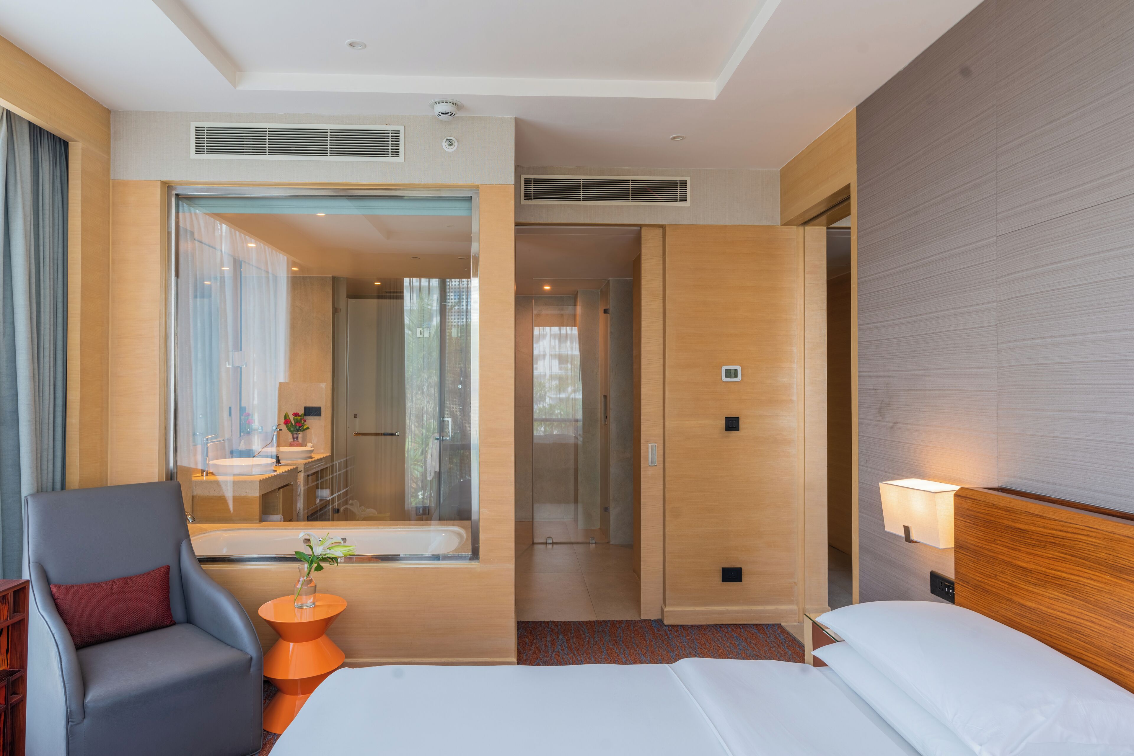A modern hotel room with a large bed, a seating area with a chair and side table, a bathroom with a bathtub, and a large window.