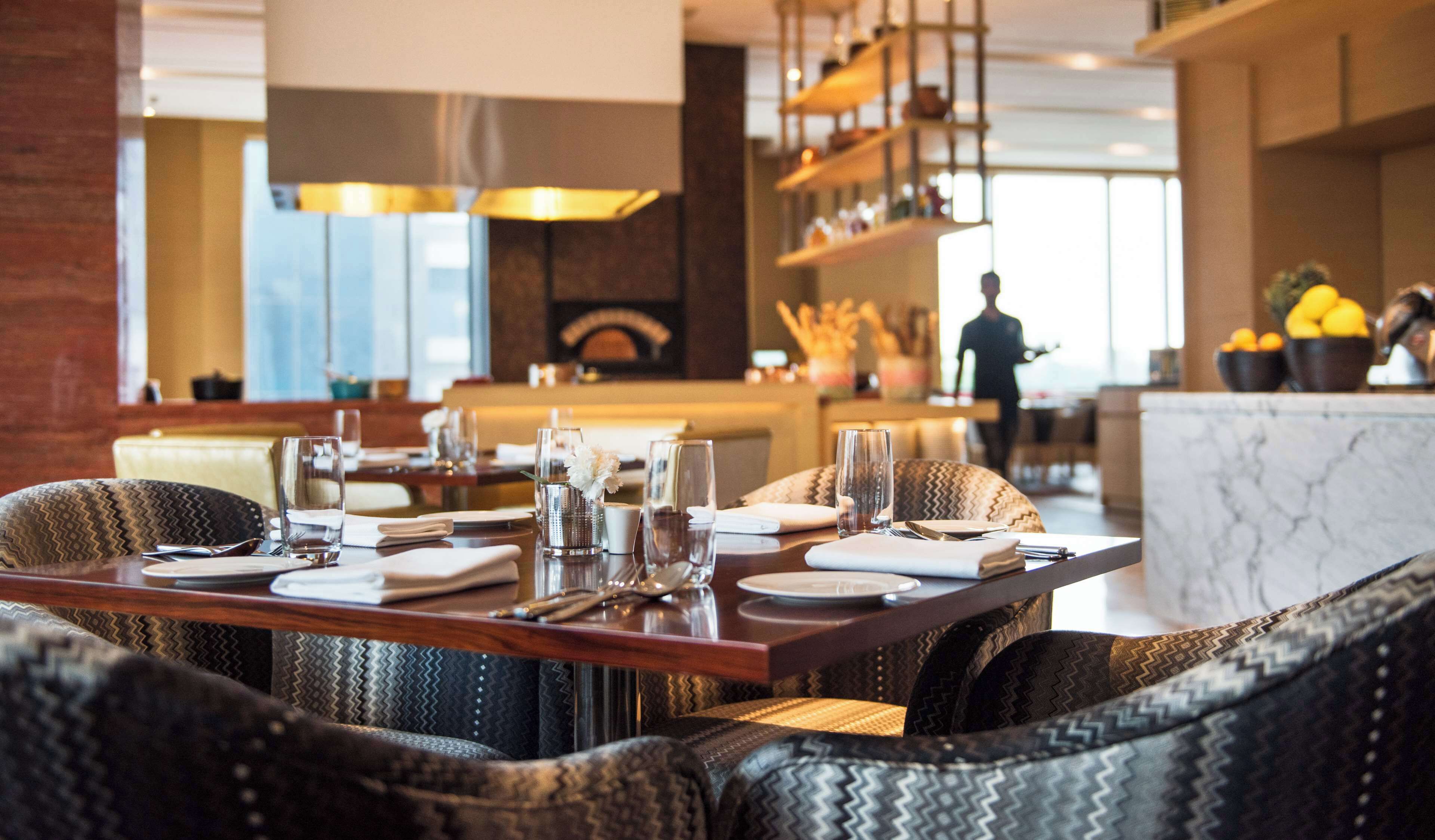 A restaurant with a modern interior, featuring a long dining table set for a meal, comfortable seating, and a view of the kitchen area.