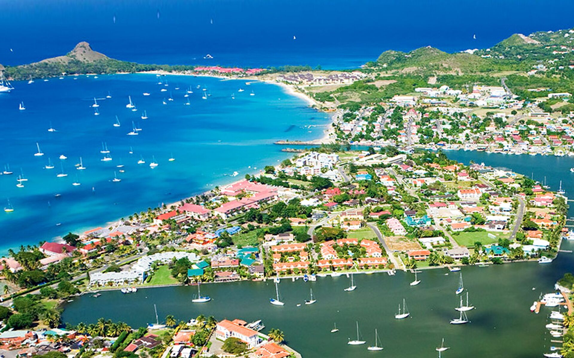 An aerial view of a coastal town with numerous boats in the water, a mix of residential and commercial buildings, and a prominent hill in the background.