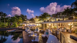 Un complexe hôtelier luxueux doté d’un espace piscine comprenant des sièges et des parasols, le tout entouré d’une végétation luxuriante et surplombant un ciel dégagé.