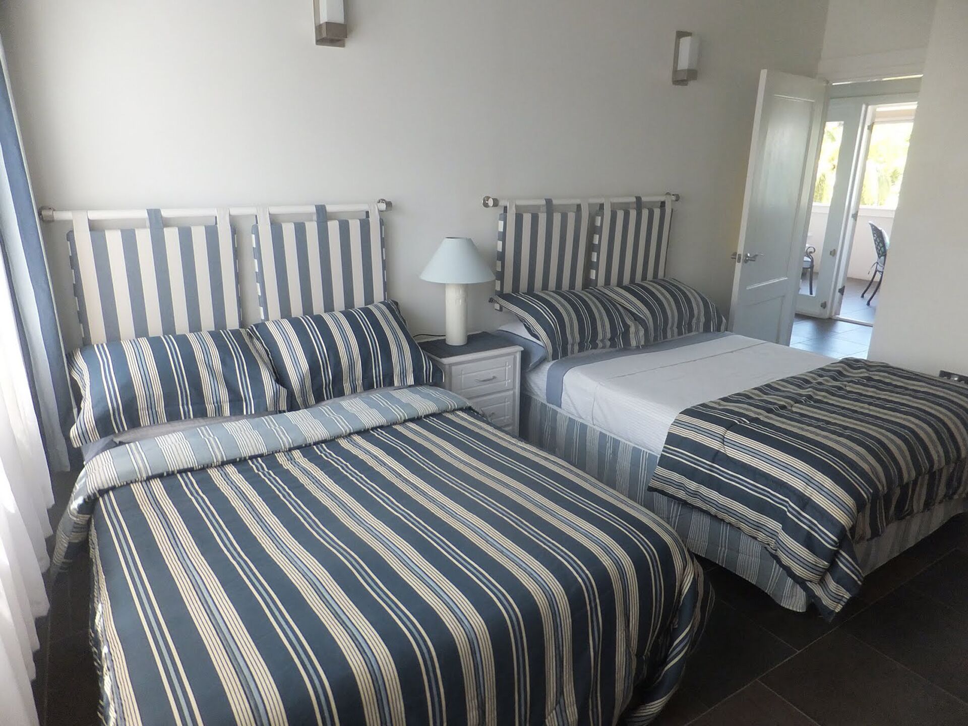 A bedroom with two beds, striped bedding, a bedside table with a lamp, and a door leading to another room.