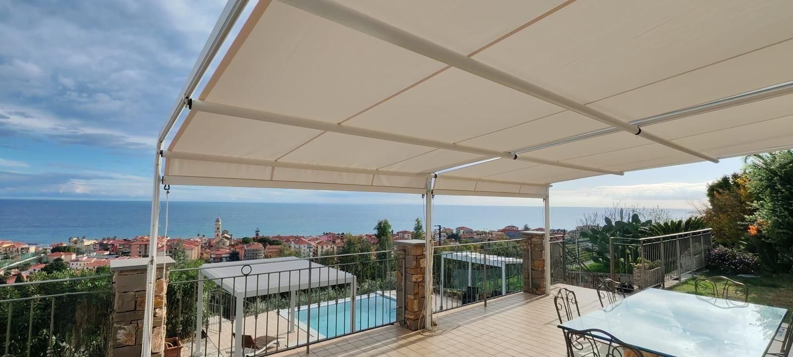 A covered outdoor area with a pool, stone pillars, and a view of the sea and town.