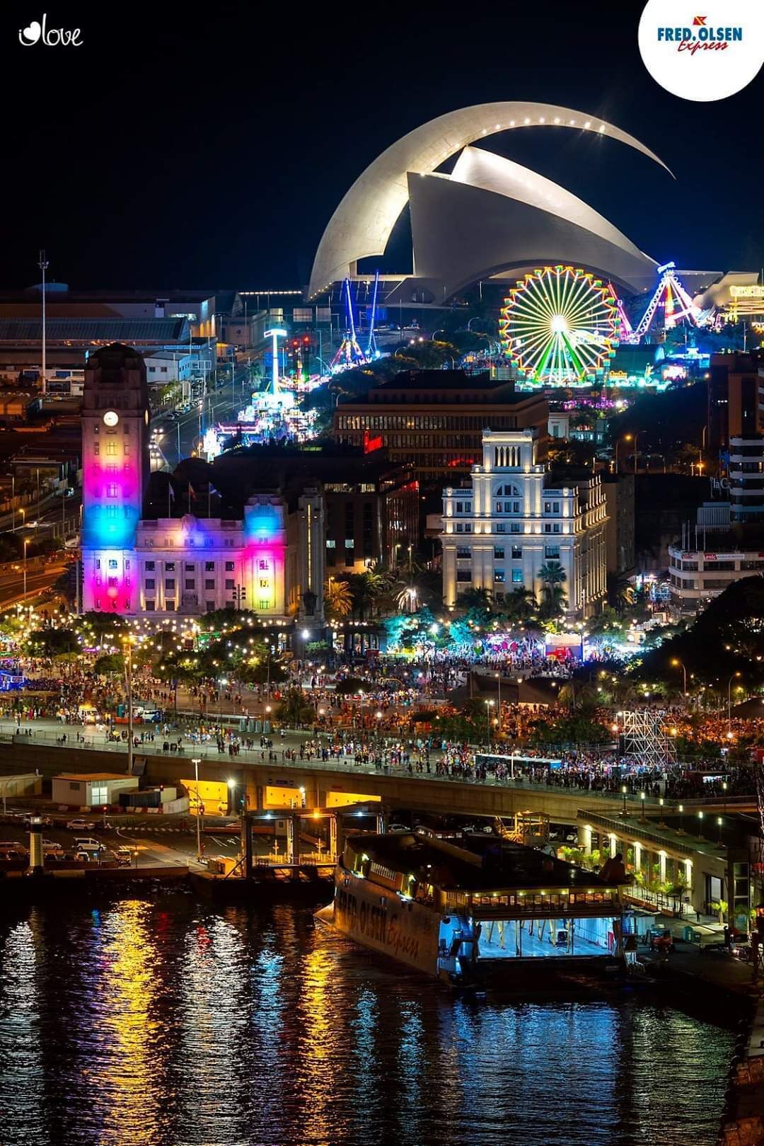 Une vue nocturne d’une ville, avec une grande roue, un bâtiment à coupole et une zone riveraine où se trouvent des bateaux.