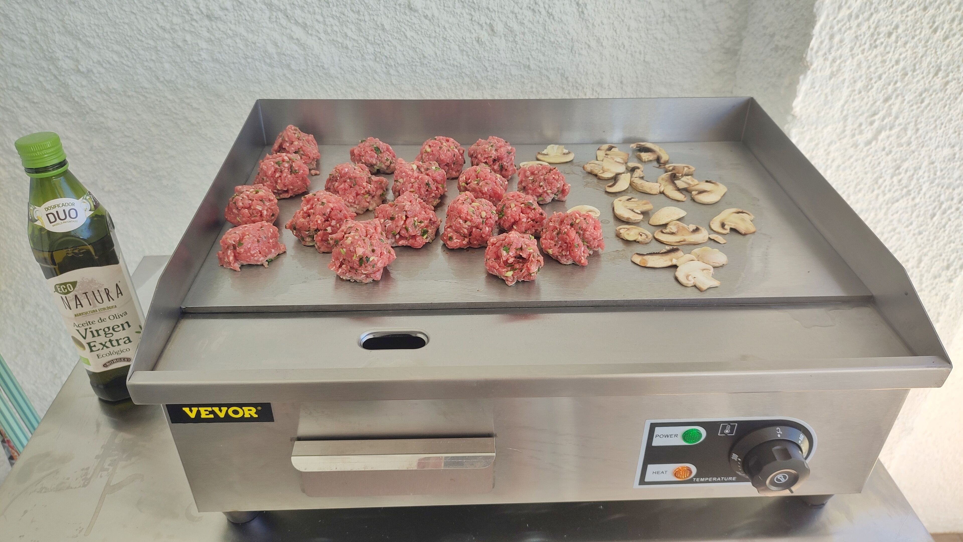 Ein Grill aus Edelstahl mit Fleisch und Pilzen, eine Flasche EVO NATURE Olivenöl sowie eine Bedieneinheit mit Einstellungen für Leistung und Temperatur.