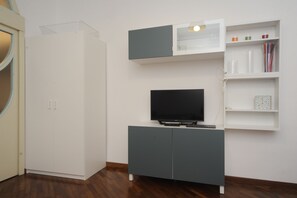 A room with a white wardrobe, a grey entertainment unit with a flat-screen TV, and a white shelf with various items.