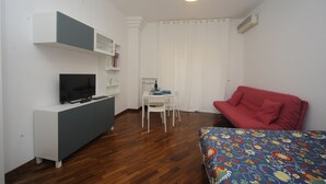 A small, well-lit room with a TV on a white cabinet, a dining table with chairs, a red sofa, and a patterned bedspread. - Jeancarlos House in the historic center (Napoli)