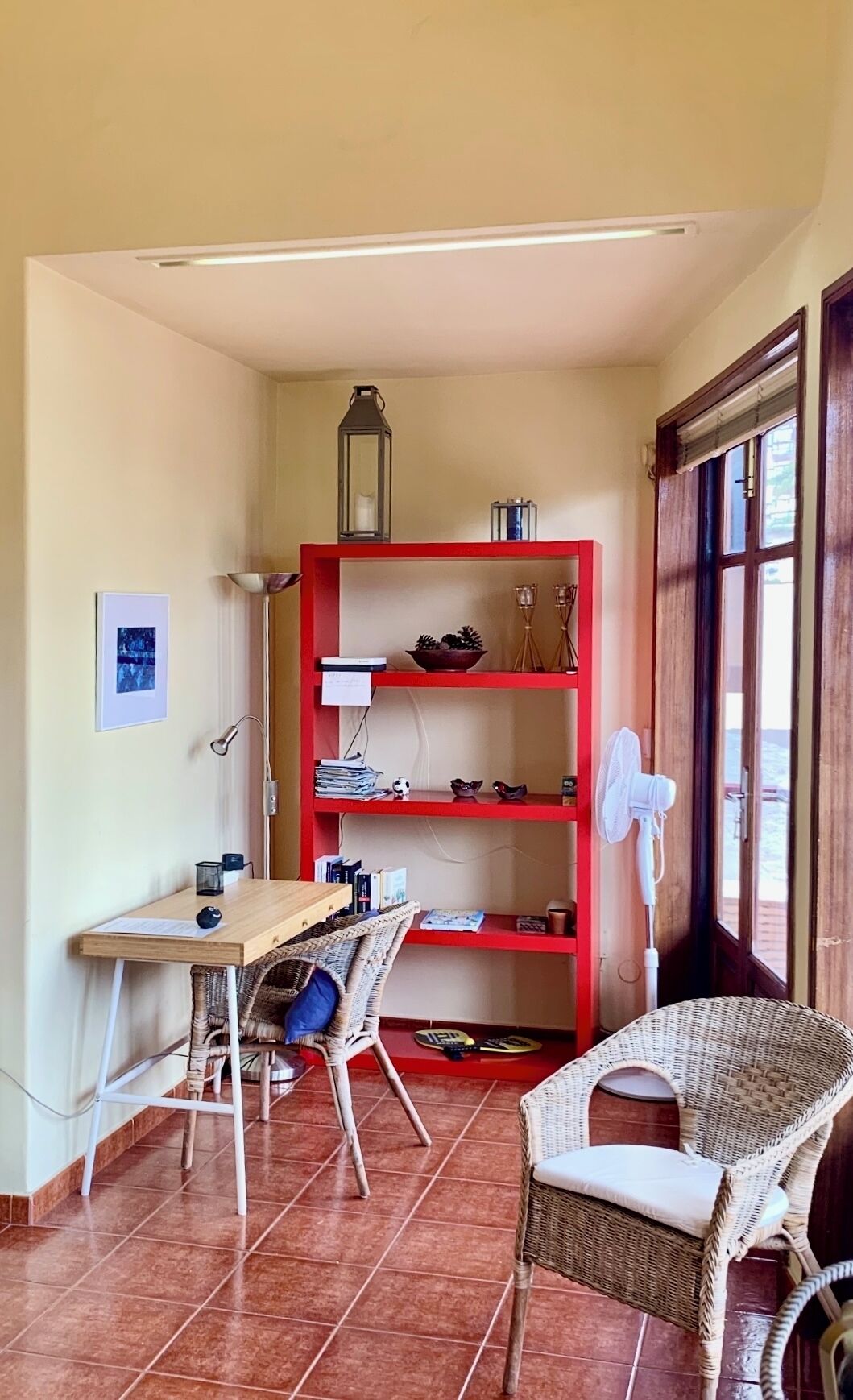 Una stanza d'angolo con una scrivania in legno, sedie in vimini, una libreria rossa e una ventola vicino alla finestra.