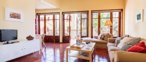 A living room with a TV, a coffee table, and a sofa. There are large windows with a view of the outside.