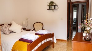 Una cama bien hecha con cabecera de madera, una mesita de noche con lámpara, un estante en la pared con una planta y un aparador de madera con un jarrón de flores.