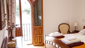Habitación de hotel con cama de madera, mesita de noche con lámpara, un florero con flores y vistas al exterior a través de una puerta de vidrio.