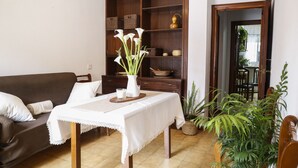 Un salón con un sofá marrón, una mesa blanca con mantel de encaje, un jarrón con flores blancas, un aparador de madera con cestas de mimbre y una planta en maceta.
