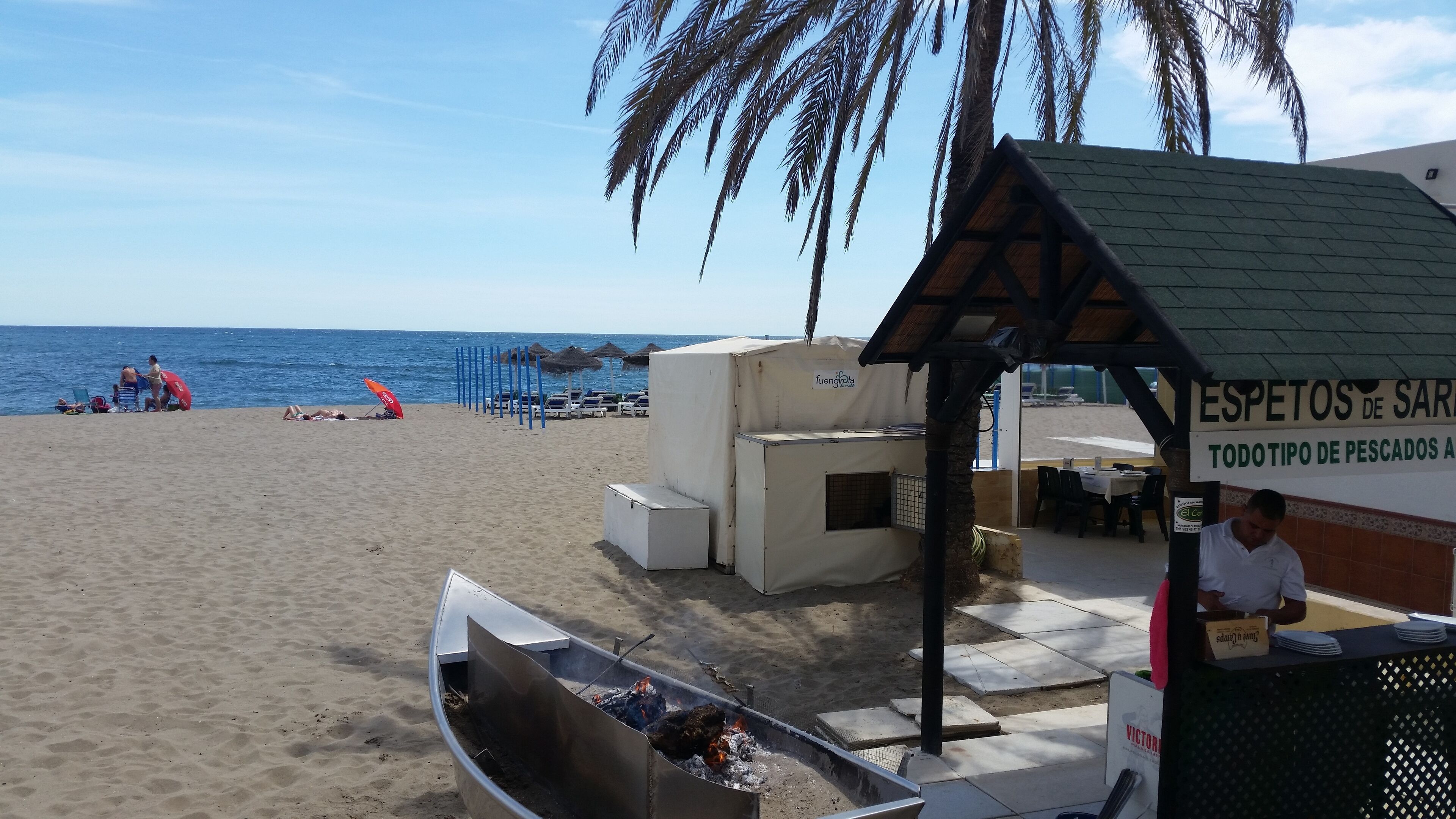 A beachside restaurant with outdoor seating, a barbecue grill, and a sign indicating seafood availability.