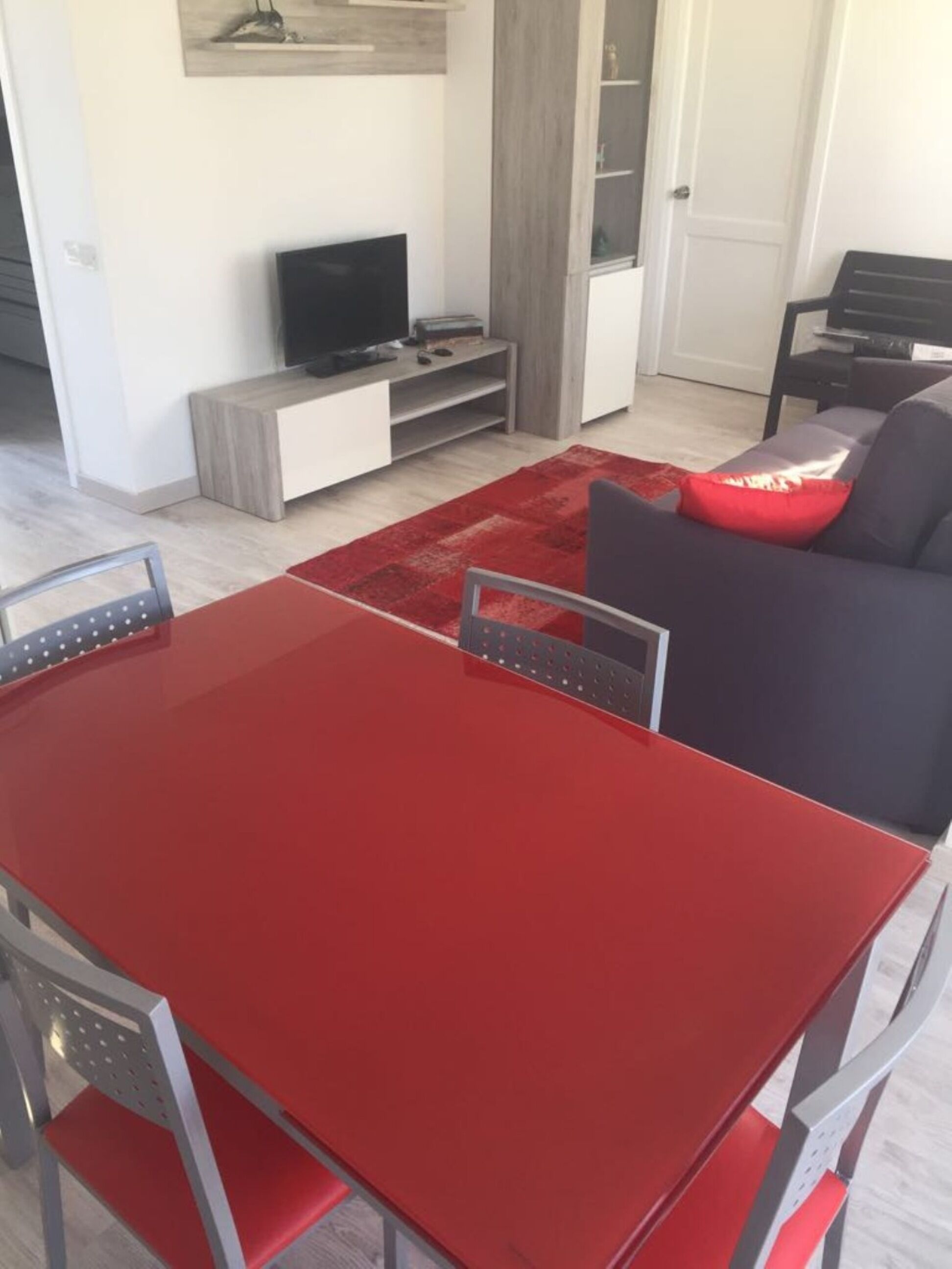 Uma sala de estar moderna com uma mesa de jantar vermelha, cadeiras cinzentas, um sofá cinza escuro com uma almofada vermelha, uma TV sobre um suporte de madeira e um tapete vermelho.