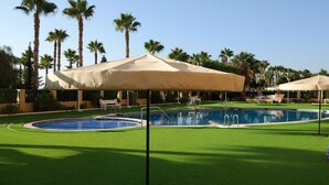 A swimming pool with a sandy edge, surrounded by green artificial grass, palm trees, and covered seating areas.
