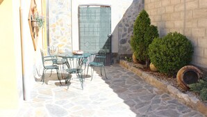 Eine Terrasse mit Steinboden, einem Tisch, Stühlen, einem Metalltor und einem kleinen Garten mit Sträuchern und einem Blumentopf.