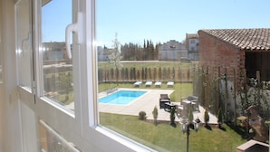 Blick aus einem Fenster auf einen Garten mit Schwimmbecken, Terrassenmöbeln und einem Backsteinhaus.