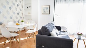 A modern living room with a grey sofa, white chairs, a small table with a plant, and a dining area with a white table and chairs.