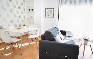 A modern living room with a grey sofa, white chairs, a small table with a plant, and a dining area with a white table and chairs.