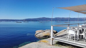 A seaside terrace with tables and chairs, overlooking a calm sea with boats and distant mountains.
