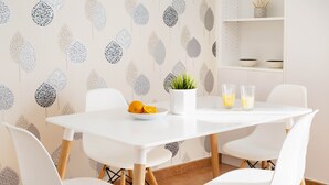 A dining area with a white table, four white chairs, a potted plant, a bowl of fruit, and two glasses of orange juice.