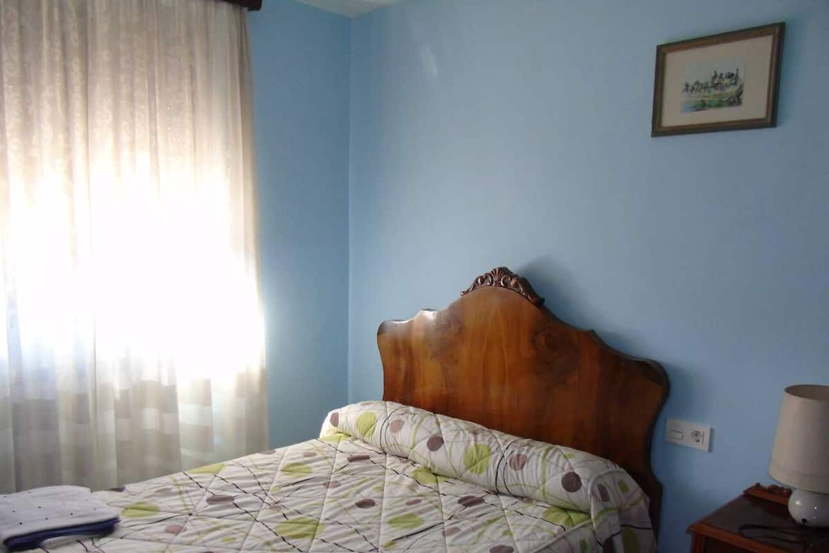 Un dormitorio con cabecera de madera, cama con colcha estampada, mesita de noche con lámpara y un cuadro enmarcado en la pared.