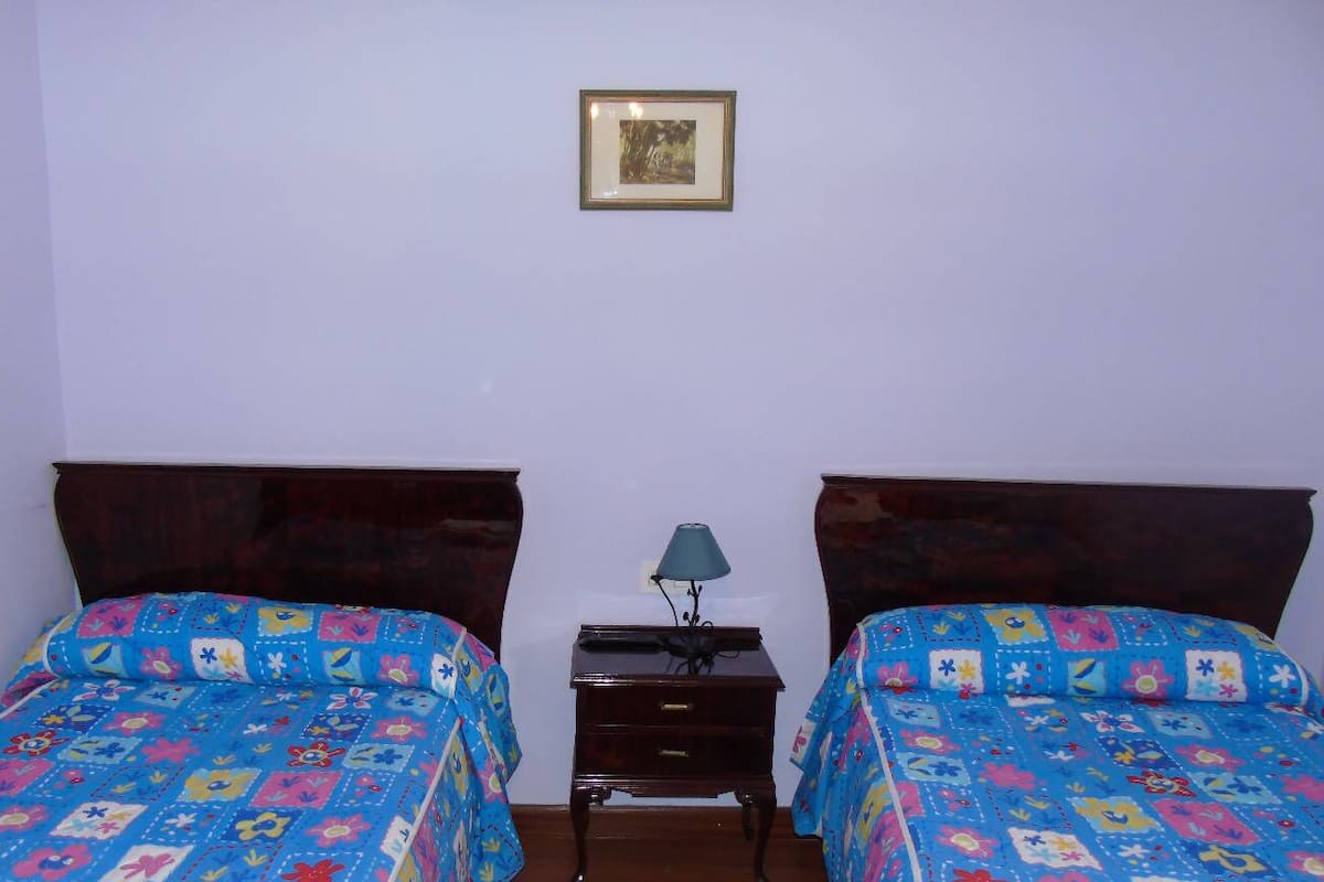 Una habitación con dos camas, cada una con una colcha floral de colores, una mesita de noche con lámpara y un cuadro en la pared.