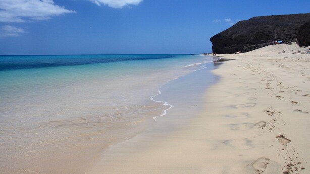 A beach with clear blue water, sandy shore, and a rocky cliff.