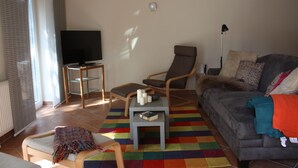 A living room with a grey sofa, a wooden chair, a small table with a book, a television, and a colorful rug. - Morning sun vacation apartment in a country house with a magnificent garden  (Westerstede)