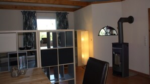 A room with a wooden ceiling, a fireplace, a dining table with chairs, and a window with curtains. - Morning sun vacation apartment in a country house with a magnificent garden  (Westerstede)