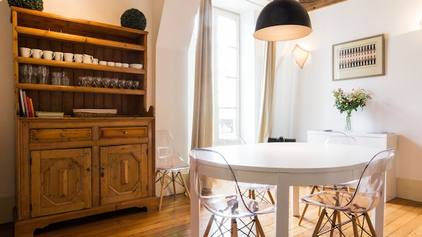 Un espace repas comprenant une table blanche, des chaises transparentes, un buffet en bois contenant de la verrerie et un luminaire suspendu.