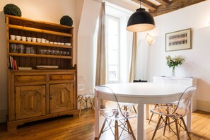 A dining area with a white table, clear chairs, a wooden cabinet with glassware, and a pendant light.