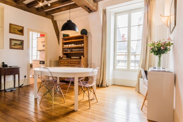 A dining area with a white table and chairs, wooden beams on the ceiling, and a window with curtains.