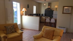 Un espace de vie compact comprenant deux fauteuils jaunes, une petite table basse, un coin cuisine avec un îlot central et une table à manger avec des chaises.