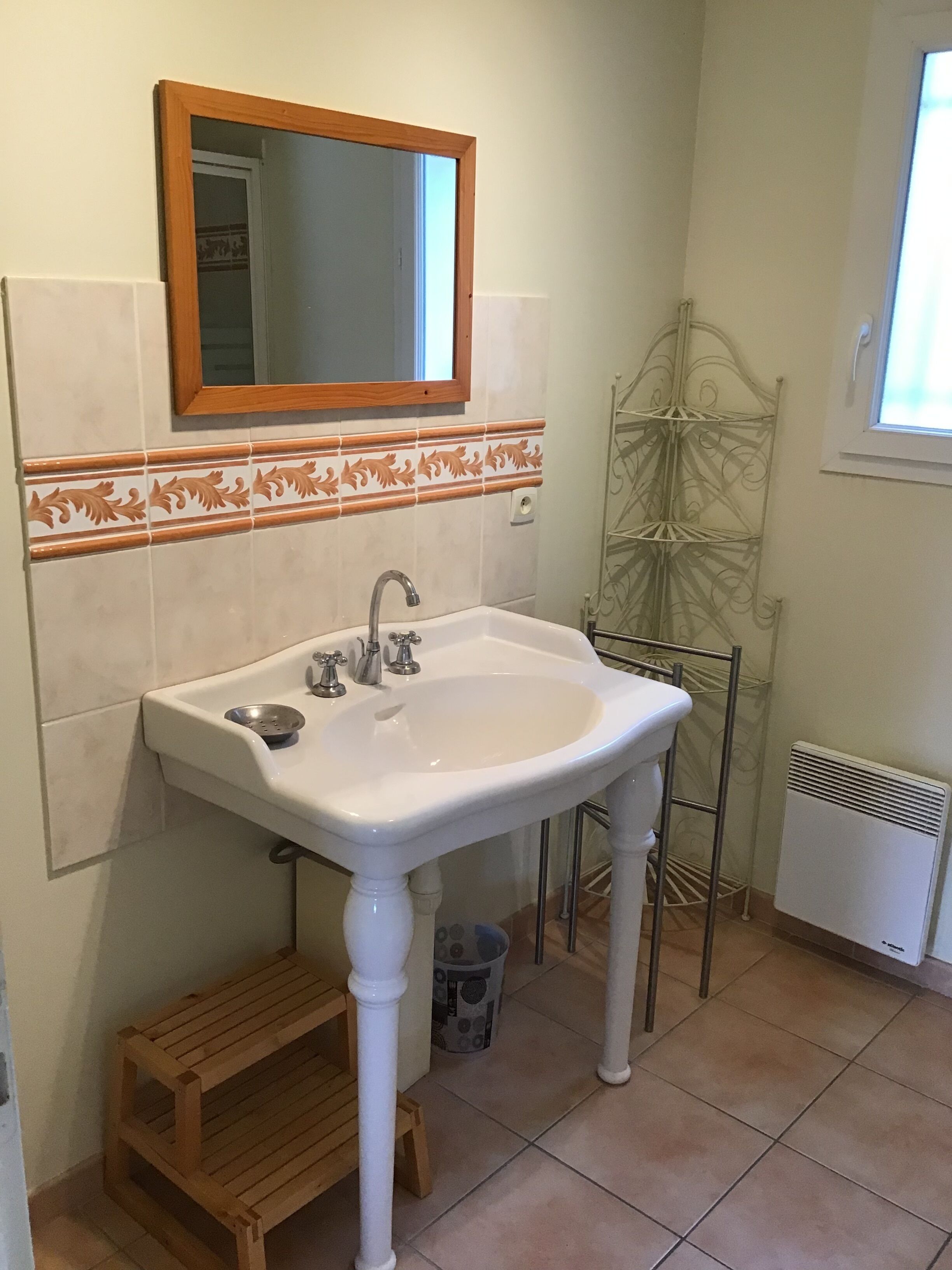 A bathroom with a white sink, a wooden stool, a metal rack, and a window.