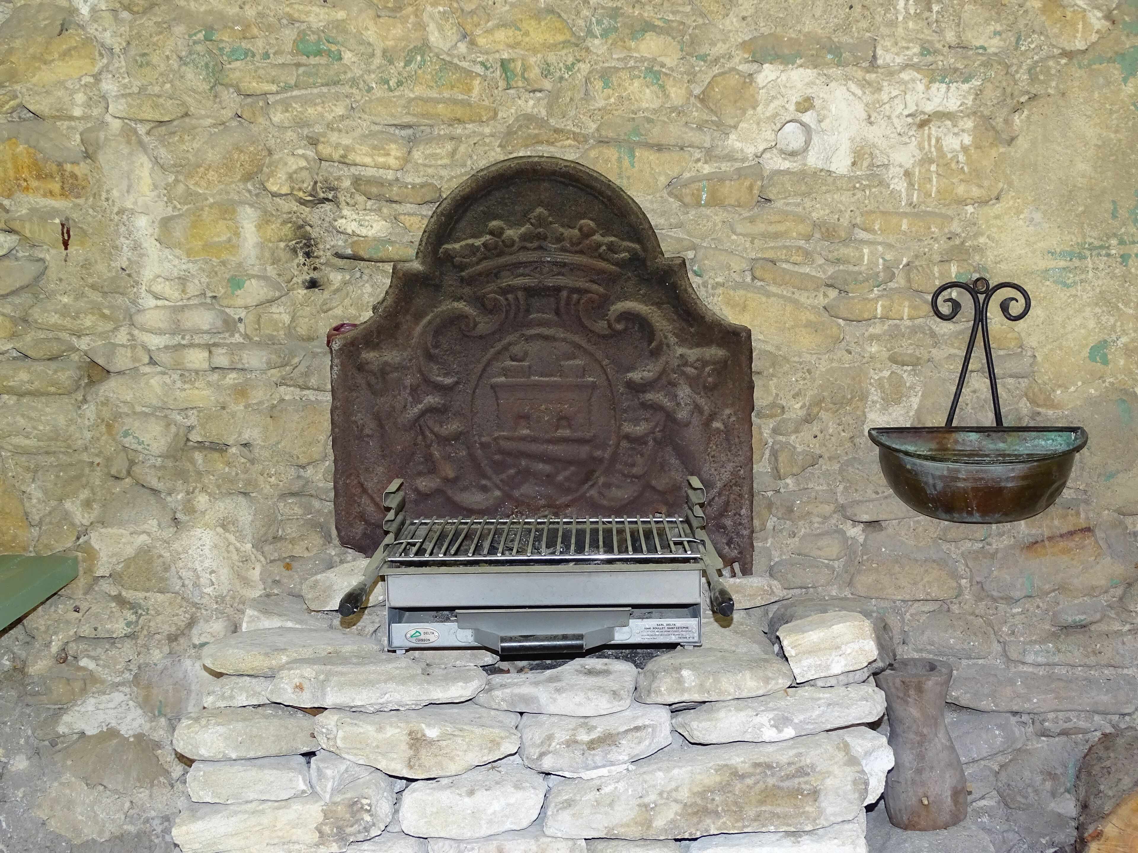 A stone fireplace with a metal grate, a decorative metal piece above it, and a hanging metal basin.