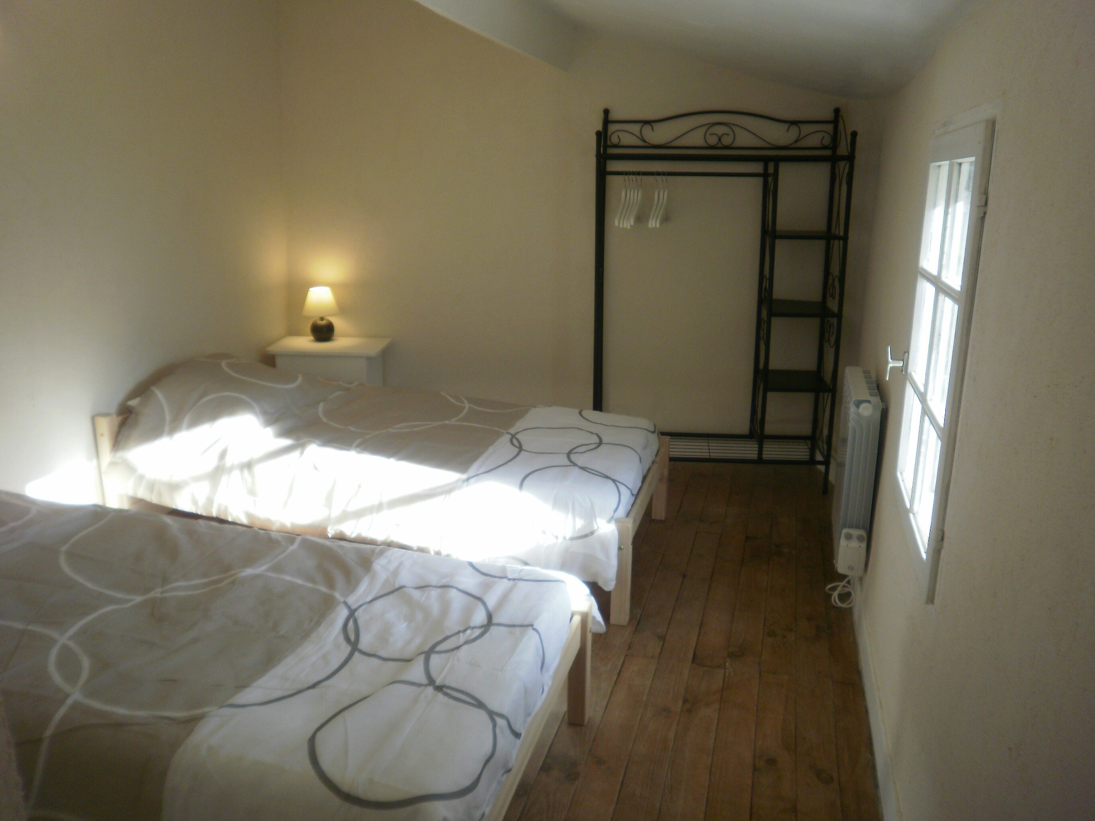 A bedroom with two beds, a nightstand with a lamp, a window with blinds, and a radiator.
