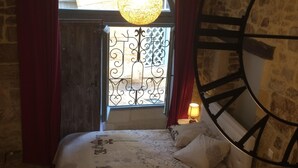 Une chambre avec un grand lit en bois, un mur en pierre, une fenêtre ornée de ferronnerie décorative et une grande horloge murale.