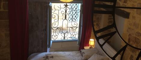 A bedroom with a large wooden bed, a stone wall, a window with decorative ironwork, and a large wall clock.