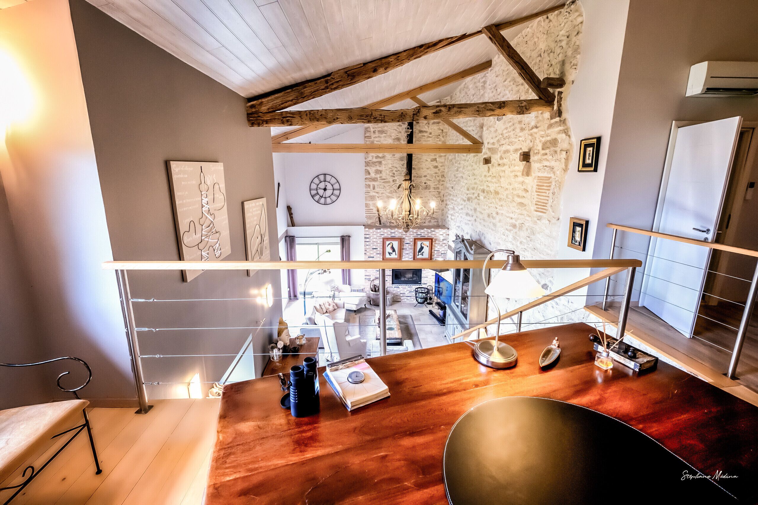 Un loft spacieux avec de hauts plafonds, des poutres apparentes, un mur en pierre et une vue sur l’espace de vie situé en contrebas.