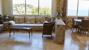 A room with a sofa, a dining table, and chairs. There are large windows with a view of the sea.