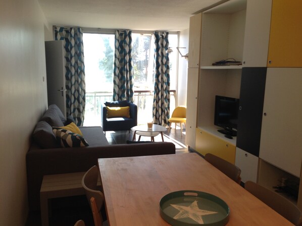 Un espacio habitable compacto con sofá, mesa de comedor, televisor y balcón con cortinas estampadas.