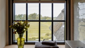 Ein Fenster mit Blick auf ein Wiesenfeld und ein Gebäude. Auf der Fensterbank steht ein Blumenvasen.
