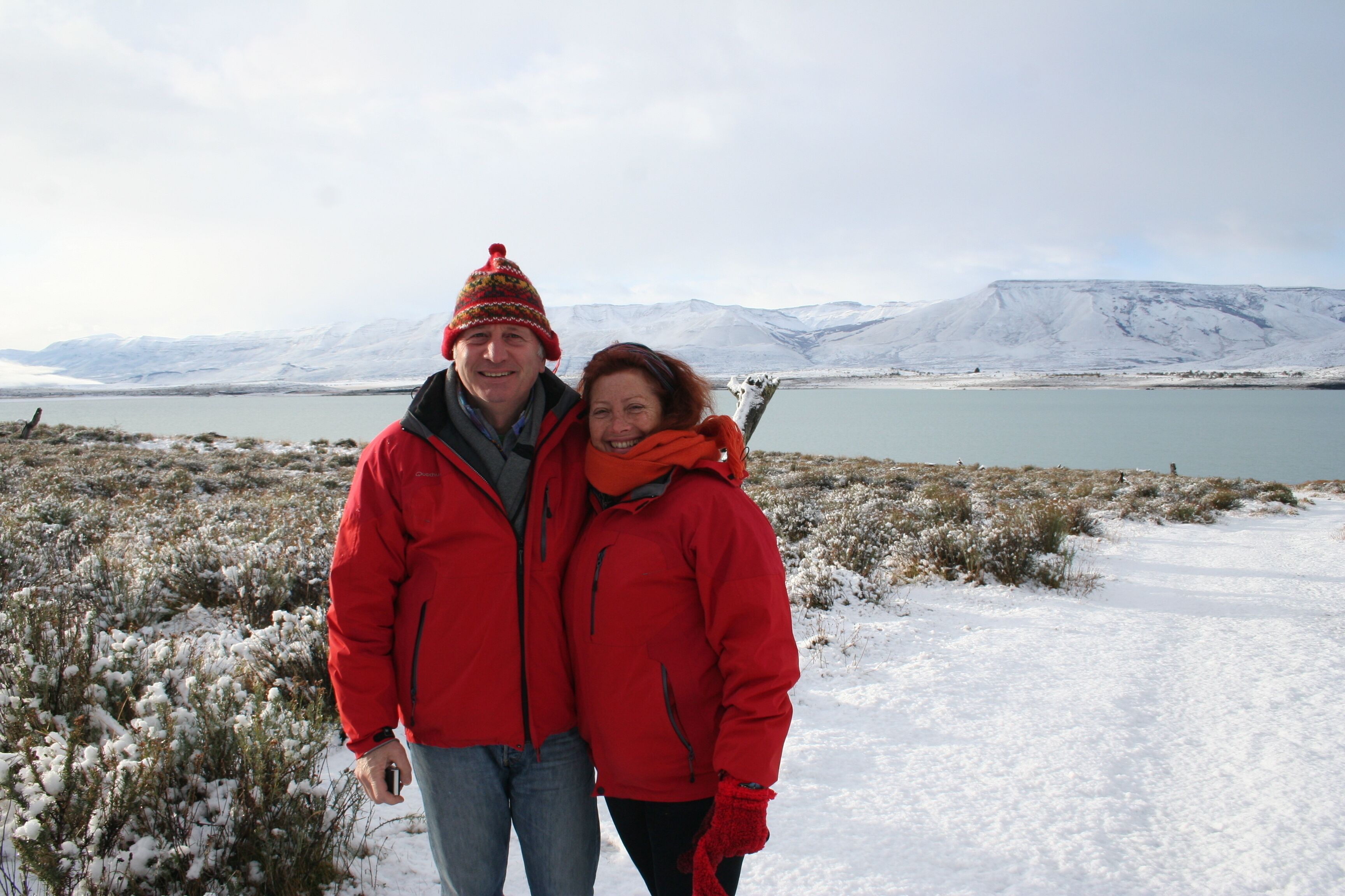 A couple in red jackets standing in a snowy landscape with a lake and mountains in the background.