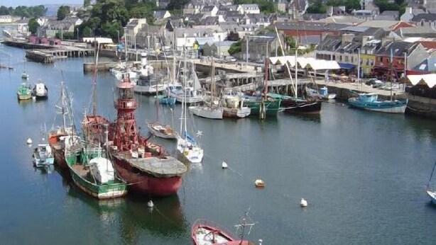 A harbor with multiple boats docked, a prominent red ship in the foreground, and a town with buildings and a church in the background. - House in a pastoral setting: 2 or 4 people (Douarnenez)