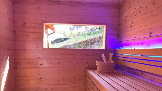 A wooden sauna with a window, a bench, a bucket, and a trash bin. - Cottage Le Meixzoté (Ferdrupt)