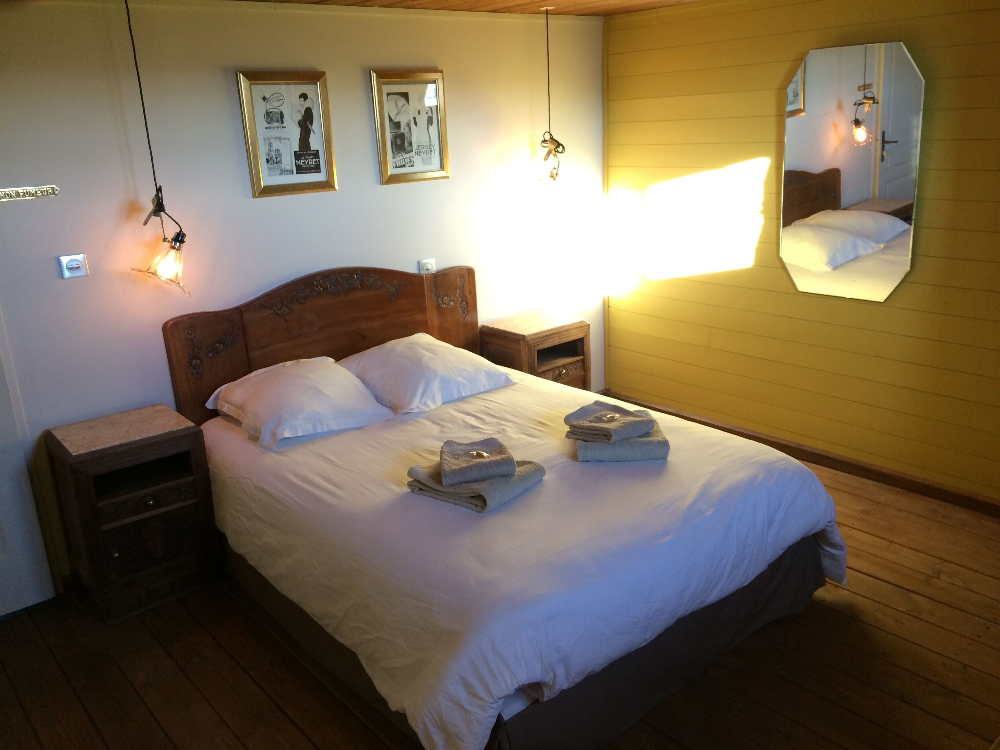Un lit bien fait, avec du linge de lit blanc, une tête de lit en bois et une table de chevet avec une lampe. Deux tableaux encadrés sont accrochés au mur, et un miroir se trouve sur le mur, à droite.